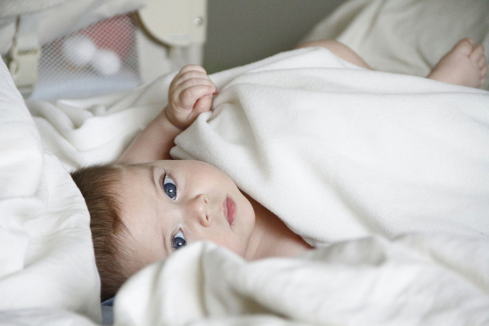 Baby peeking from under a white blanket, with blue eyes and soft expression.