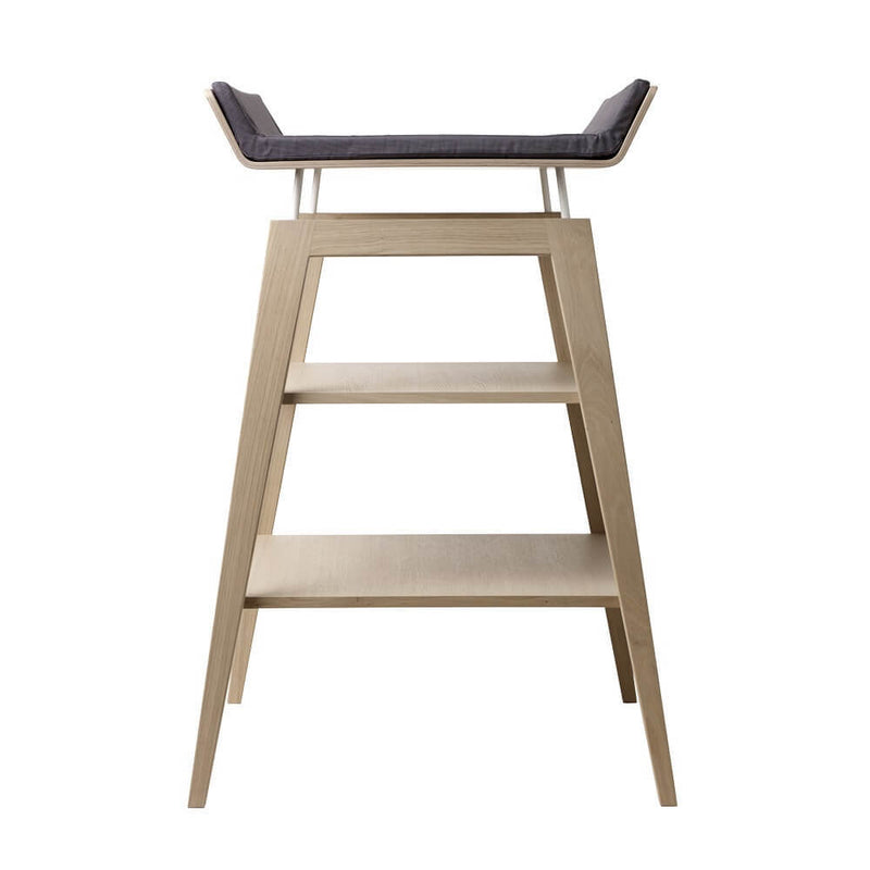 The Leander Linea Changing Table by Leander features angled legs, a light wood finish, two lower shelves inspired by baby changing stations, and a dark cushioned top.