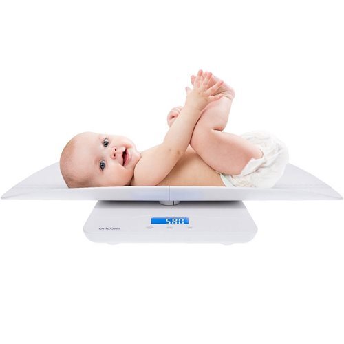 A baby in a diaper lies on its back holding its feet on an Oricom 1100 Digital Baby Scale by Oricom, with the weight shown in blue digits against a plain white background.