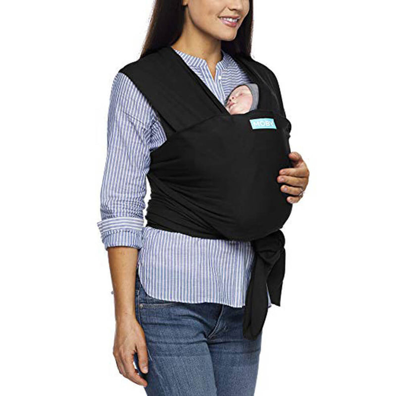 A woman in a striped shirt and jeans carries a sleeping baby in a black Moby Evolution Wrap by Moby, the soft fabric baby carrier tied securely at her waist.