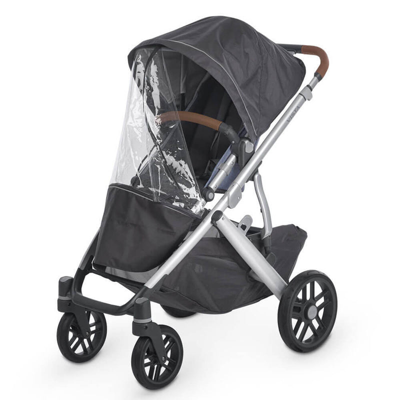The UPPAbaby Cruz/Vista Rain Shield by UPPAbaby is a black stroller with a transparent rain cover, silver frame, four wheels, storage basket underneath, and brown handles.
