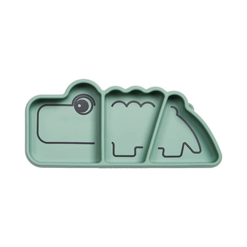The Done By Deer Silicone Stick & Stay Snack Plate Croco is a green, sectioned suction baby plate by Done By Deer, shaped like a crocodile with black line details for eyes, scales, and mouth dividing three compartments.