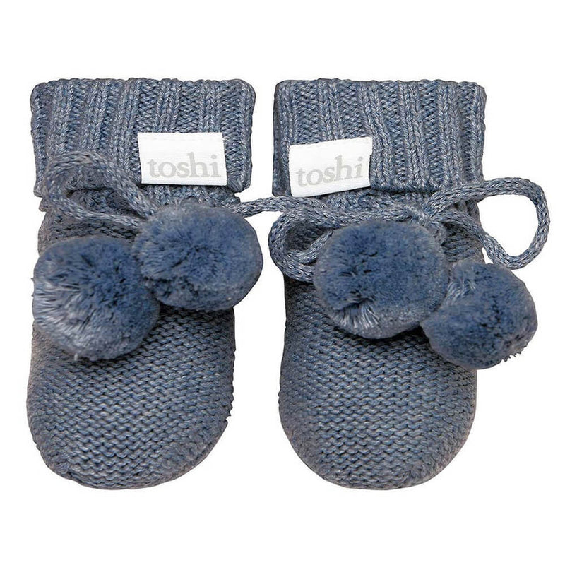 Toshi Booties by Toshi—knitted blue organic baby booties with folded cuffs, pom-poms, and branded labels on each cuff—are perfect for a thoughtful newborn gift box.