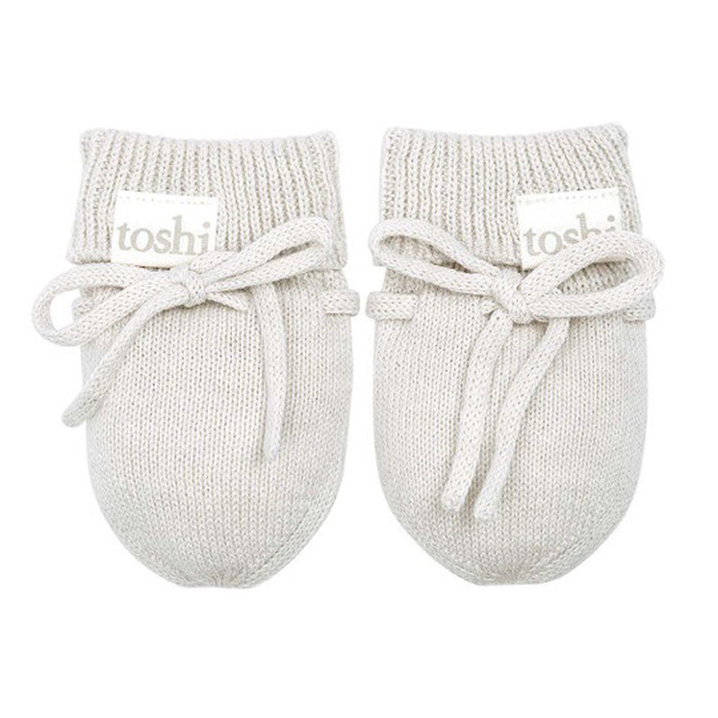 Toshi Organic Mittens Marley: A pair of soft, knitted, light beige organic cotton baby mittens with ribbed cuffs and tied bows. Each features a "toshi" tag. Displayed side by side on a white background. Brand: Toshi.