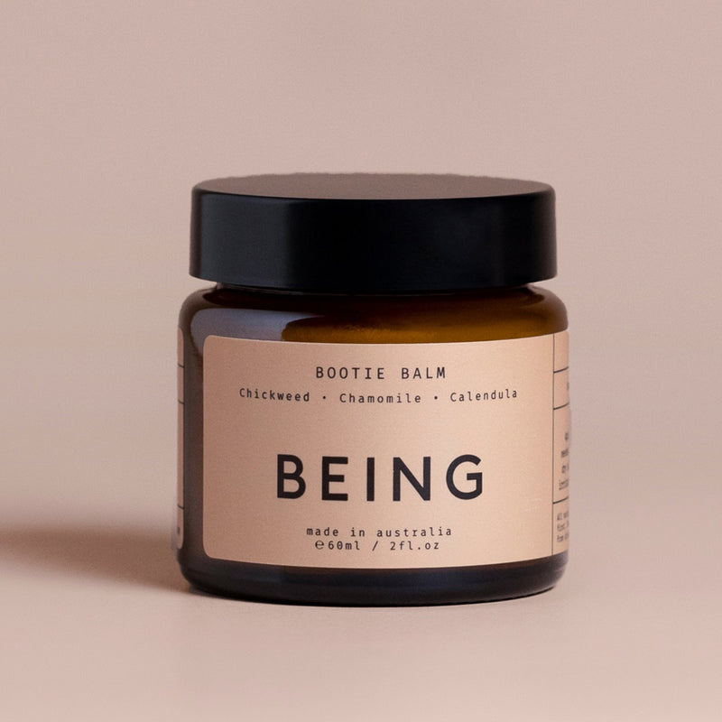 A small amber jar with black lid labeled "Being Skincare Bootie Balm" features chickweed, chamomile, and calendula. This 60ml natural baby balm from Being Skincare is made in Australia to soothe delicate baby skin, set against a soft beige background.