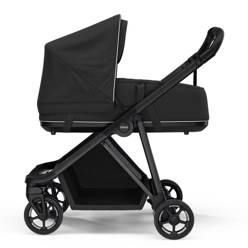 The Thule Shine Stroller + Thule Bassinet by Thule is a sleek black city pram with four wheels, a roomy storage basket, large canopy, and bassinet attachment, shown from the side against a white background.