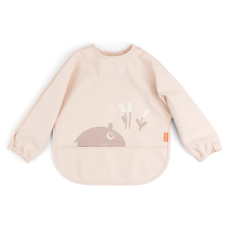 The Done By Deer Sleeved Pocket Bib Ozzo Powder is a light beige, long-sleeve adjustable baby bib with elastic cuffs and a bottom pocket. It features a simple brown hippo and cattails illustration on the front.