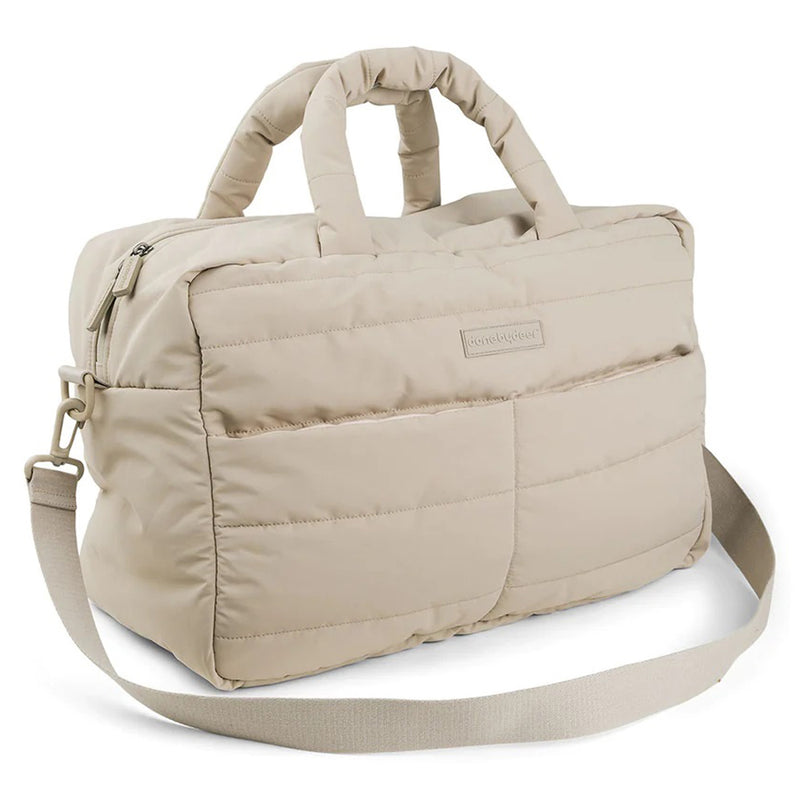 The Done By Deer Quilted Changing Bag Sand by Done By Deer is a beige recycled polyester bag with two padded handles, a zippered top, two exterior pockets, and an adjustable shoulder strap. It features a plain, modern quilted design.