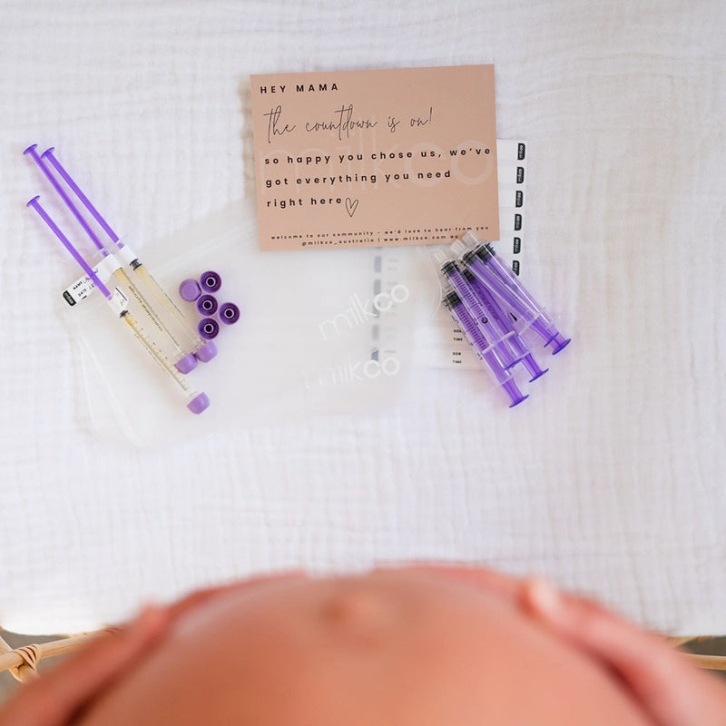 A top-down view of the Milkco Mini Colostrum Kit by Milkco, including a sterile syringe and accessories for colostrum collection, arranged on a white surface with the blurred outline of a pregnant belly visible at the bottom.