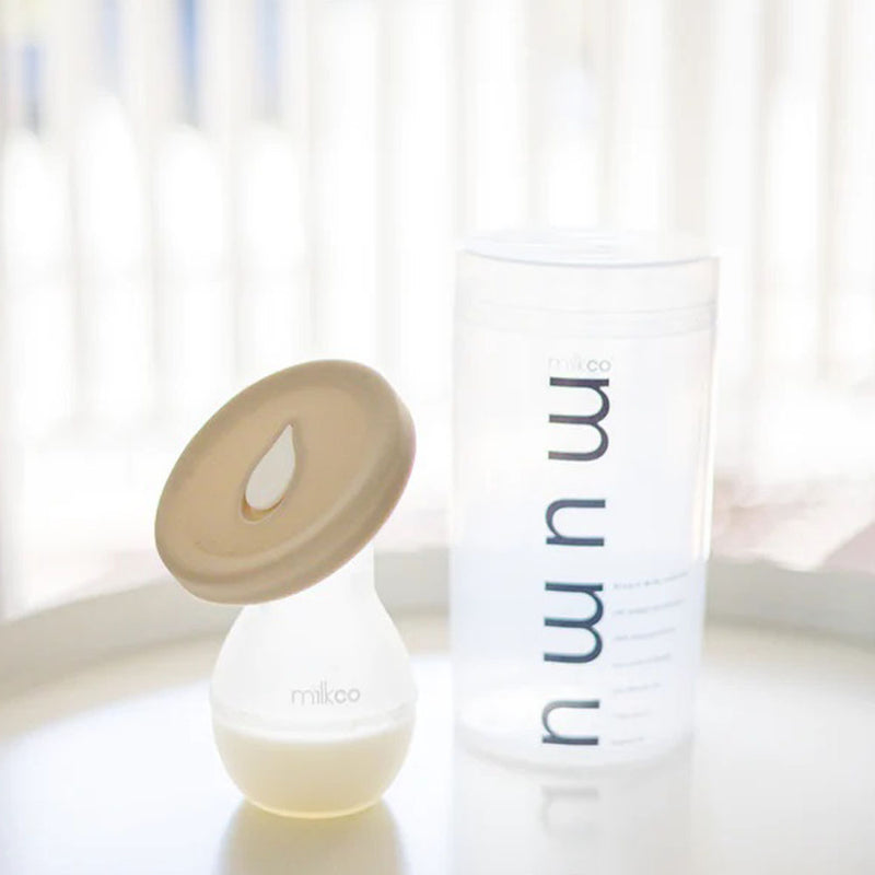 The Milkco Mumu Breast Milk Collector with a beige rim sits beside a transparent “mum”-labeled storage container on a white surface, softly lit by natural light.