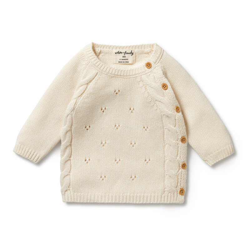 The Wilson & Frenchy Ecru Knitted Pointelle Kimono Cardigan is a cream, long-sleeve newborn cardigan with a diamond knit pattern, side cable detailing, and four brown buttons. Made from a cozy cotton bamboo wool blend.