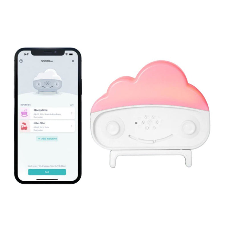 A smartphone displaying an app rests beside the Happiest Baby Snoobie White, a small cloud-shaped smart soother with a pink top, smiling face, buttons, and built-in white noise machine features, standing upright on its stand.