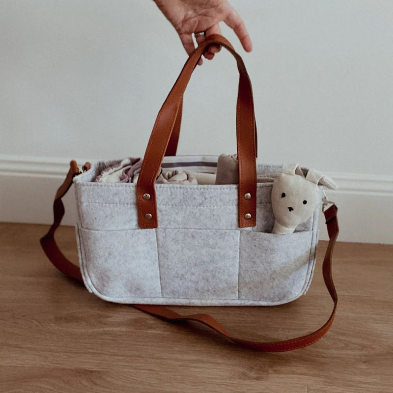 A hand holds the Angus The Label Nappy Caddy Grey with vegan leather straps on a wooden floor. A soft toy bear peeks from a side pocket, making this Angus The Label organiser both stylish and practical for any nursery.