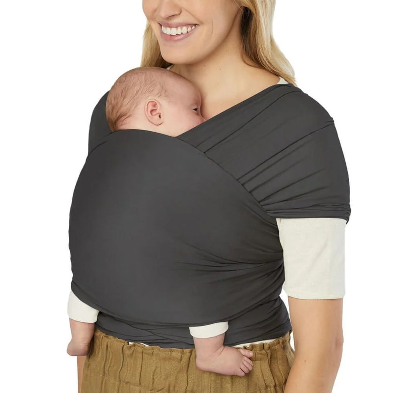 A smiling woman wears the ErgoBaby Aura Sustainably Sourced Knit Baby Wrap Soft Black, holding a sleeping infant close to her chest. The baby is snug and secure in the soft, sustainably sourced ergonomic wrap.