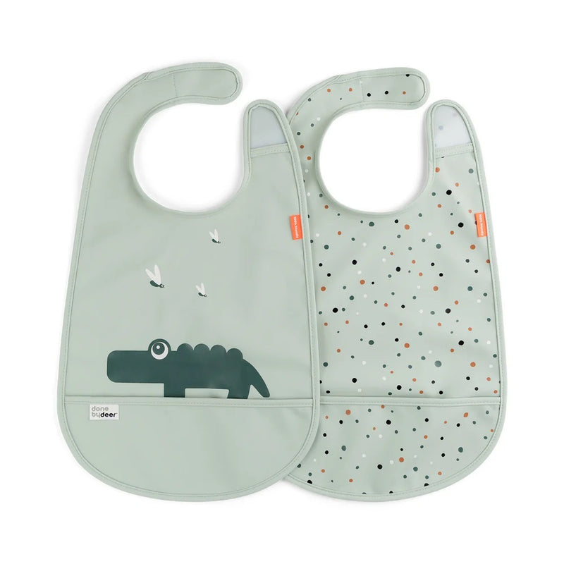 The Done By Deer Bib W/Velcro 2-Pack Croco Green includes two waterproof bibs: one with a crocodile and birds, and one with multicolored dots. Both have bottom pockets and adjustable neck straps.