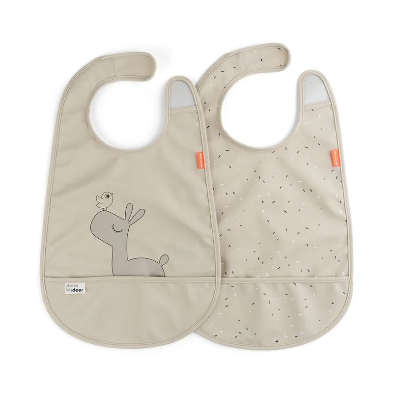 The Done By Deer Bib W/Velcro 2-Pack Lalee Sand includes two beige bibs: one with a smiling deer and bird, the other with black speckles. Both feature velcro closures, orange side tags, and are by Done By Deer.