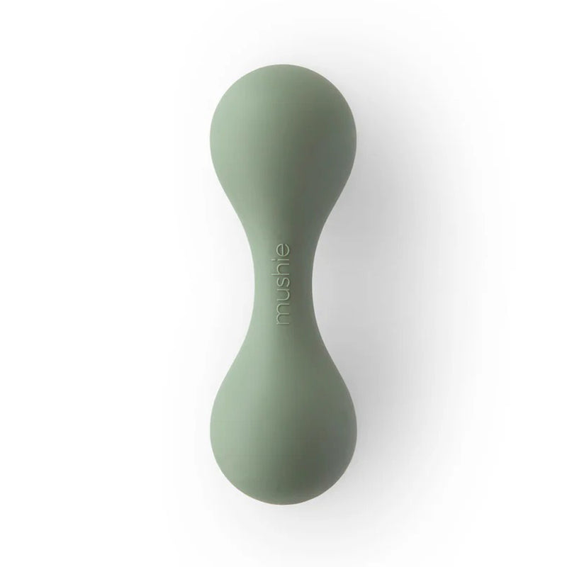 The Mushie Silicone Baby Rattle Toy Dried Thyme is an hourglass-shaped, light green rattle made from food-grade silicone with a smooth finish and "mushie" printed in white on the center. Ideal as a sensory toy for babies.