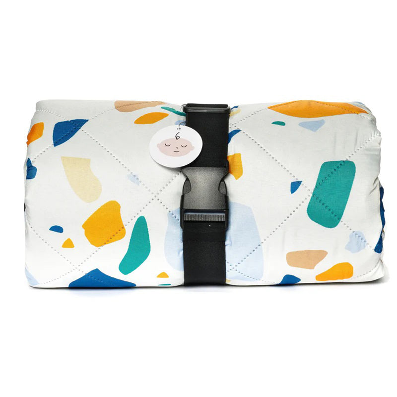 The IZIMINI Charlie Picnic Rug is a folded, quilted mat with a white background and colorful abstract shapes in yellow, blue, green, and orange. It’s water-resistant and fastened by a black strap with a round pink tag.