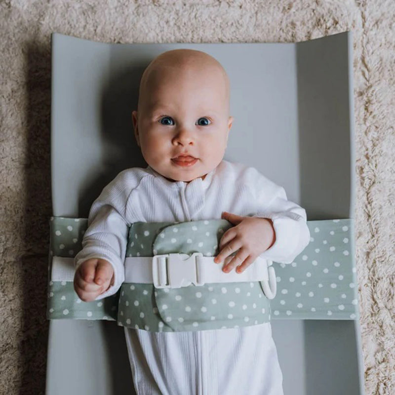 A baby in a white onesie lies on a gray changing pad wearing the WriggleBum Wrigglebum Nappy Changing Harness Sage Whimsy—ideal for quick nappy changes. The baby gazes up with bright blue eyes and a slight smile.