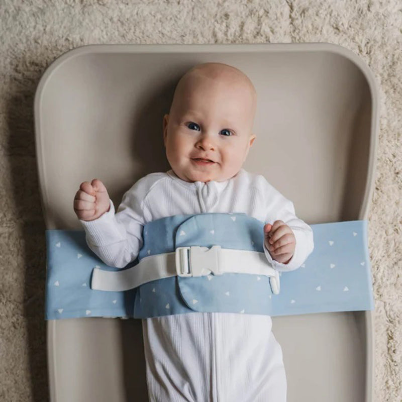 A smiling baby in a white onesie lies on the WriggleBum Wrigglebum Nappy Changing Harness Breezy Blue, secured with a blue strap featuring white hearts and a white buckle, on a beige surface.