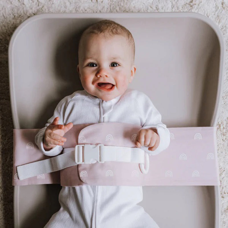 A smiling baby in the WriggleBum Nappy Changing Harness Rainbow Blush, secured with a white buckle for quick, easy nappy changes. Perfect for portable baby changing at home or on the go.
