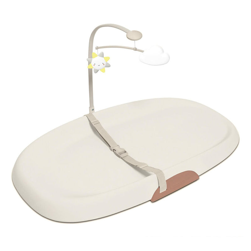 The Skip Hop Wipe Clean Changing Pad Oat by Skip Hop is a beige, contoured pad with safety straps and an overhead mobile featuring a smiling sun and cloud, all set against a white background.