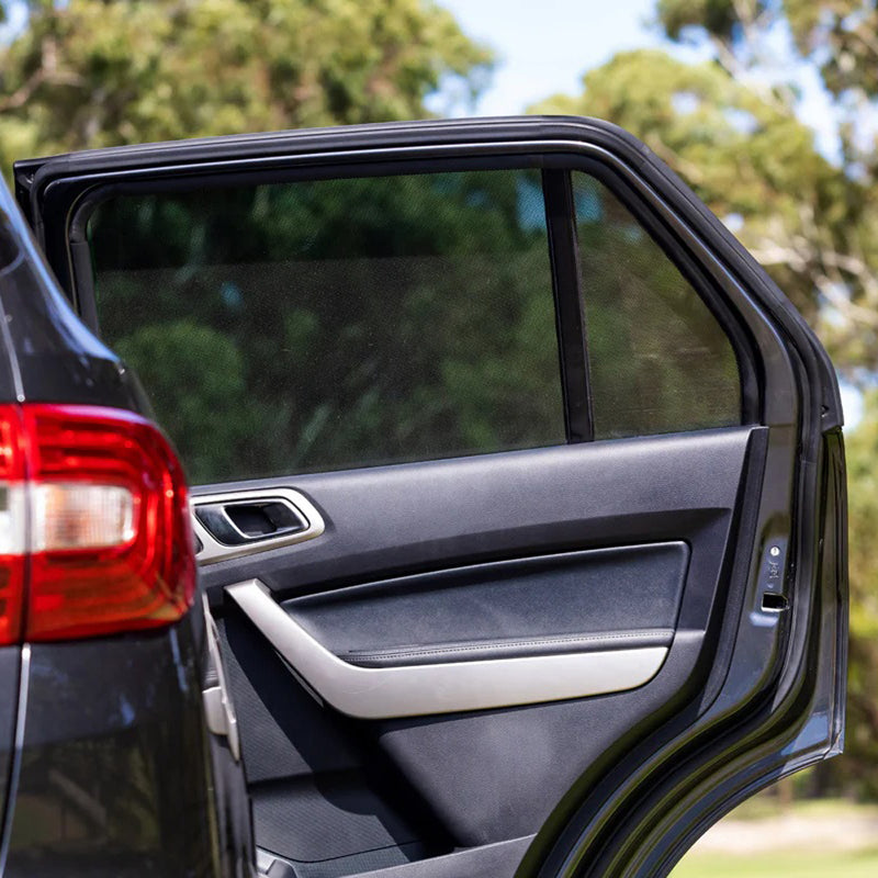 A close-up of a black car with the rear passenger door open reveals the interior fitted with Toddler Tints window shades by Toddler Tints, providing sun protection for kids. Blurred trees and greenery are visible in the background.