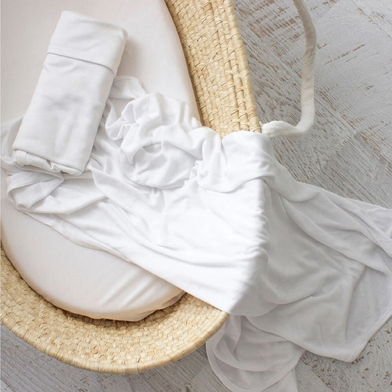 The Bubba Blue Bamboo Jersey Wrap from Bubba Blue, paired with a white fitted sheet, is draped over a woven Moses basket on a light wooden floor, creating a cozy and inviting setting for your baby.