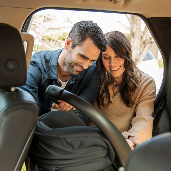 A smiling couple uses the Baby Village Car Seat Fitting Service by Baby Village, adjusting an infant seat together in a sunlit car, looking happy and focused during the installation.