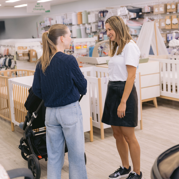 Two women talk near a crib display in Baby Village, one with a stroller, enjoying the Baby Village VIP Personal Shopping Experience and expert baby product advice surrounded by shelves of baby products and accessories.