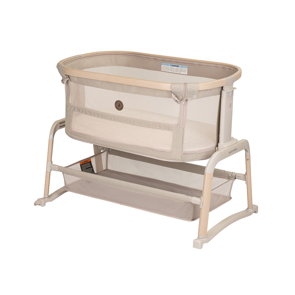 The Maxi Cosi Iora Air Lux Bedside Sleeper by Maxi Cosi is a beige and light wood baby bassinet with breathable mesh sides, an extra-comfy mattress, an open storage shelf below, and a sturdy frame, shown against a white background.