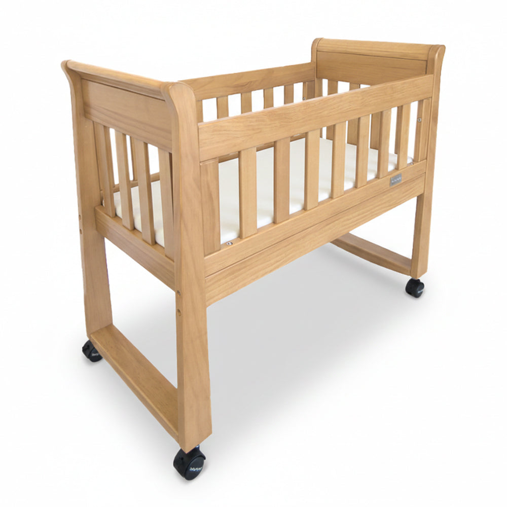 The Babyhood Sandton Sleigh Cradle by Babyhood features slatted wooden sides, a flat base, and four black caster wheels, shown against a white background.