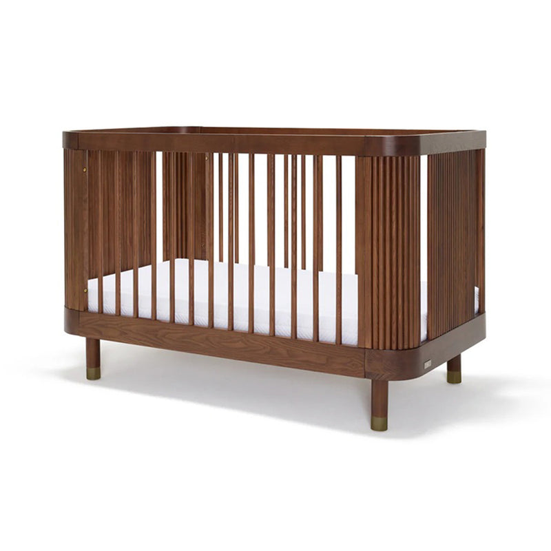 The BabyRest Juno Cot Luxe Walnut is a modern wooden crib with vertical slats, rounded corners, and a fluted facade. Made from European Ash in a dark brown finish, it features short legs with light tips and includes a white mattress.