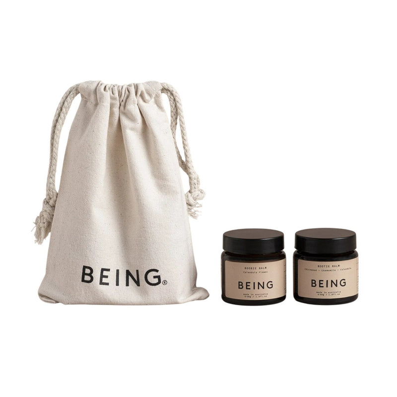 A beige drawstring bag labeled "BEING" sits beside two small jars with beige labels and black lids. The Being Skincare Mother & Baby Set is ideal for use as a nappy rash balm or postpartum balm.