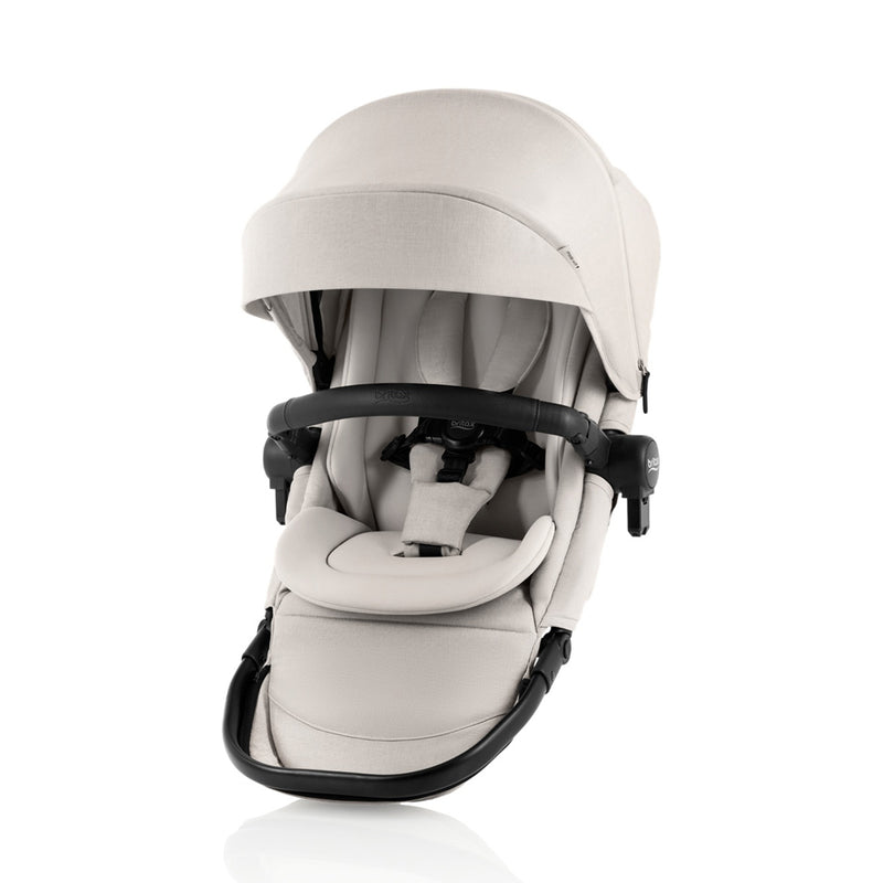 The Britax Safe-n-Sound Aptana 2nd Seat is a beige baby stroller seat with canopy, safety harness, and black bumper bar—newborn ready and designed for double stroller use. Shown against a white background.