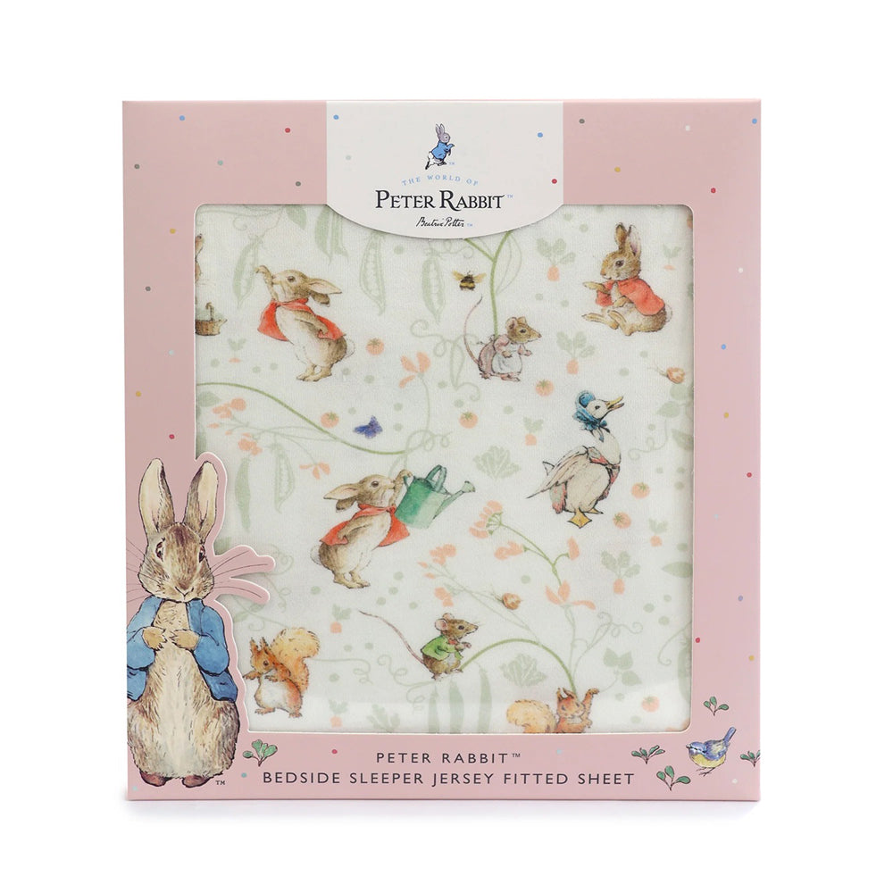 The Bubba Blue Flopsy Mopsy & Cottontail Bedside Sleeper Fitted Sheet features charming illustrations on soft jersey fabric. Packaged in a pink box with a clear window, decorative artwork, and product details at the bottom.