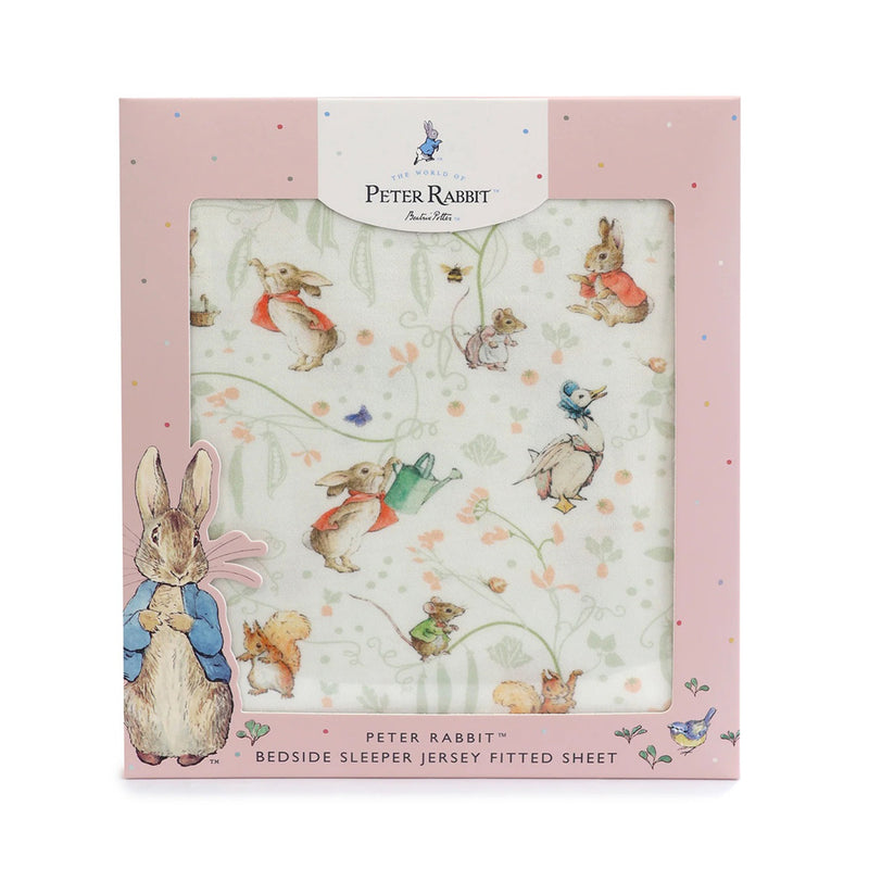 The Bubba Blue Flopsy Mopsy & Cottontail Bedside Sleeper Fitted Sheet features charming illustrations on soft jersey fabric. Packaged in a pink box with a clear window, decorative artwork, and product details at the bottom.