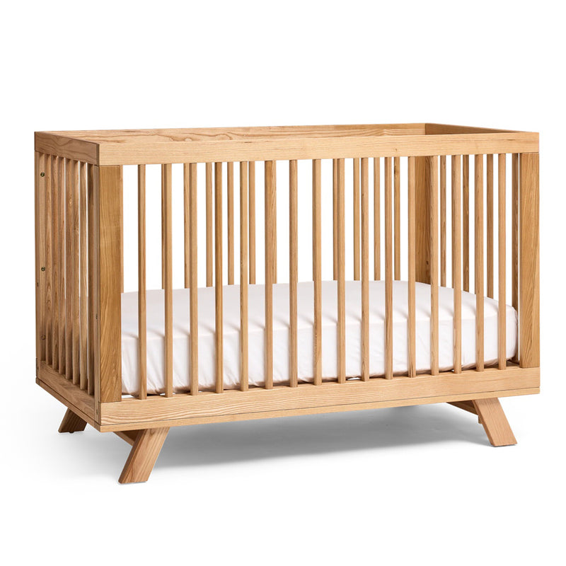 The Cariboo MacKenzie Cot by Cariboo is a modern Ash timber crib with vertical slats, angled legs, and a white mattress, shown on a plain white background. It easily converts into a toddler bed.