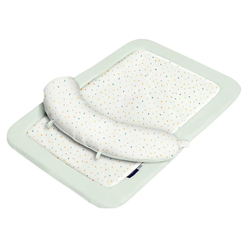 The ClevaMama Newborn Tummy Time Mat by Clevamama is a soft, rectangular mat with a light green border, white multicolored dotted center, and a detachable crescent-shaped ClevaFoam pillow to help prevent flat head syndrome.