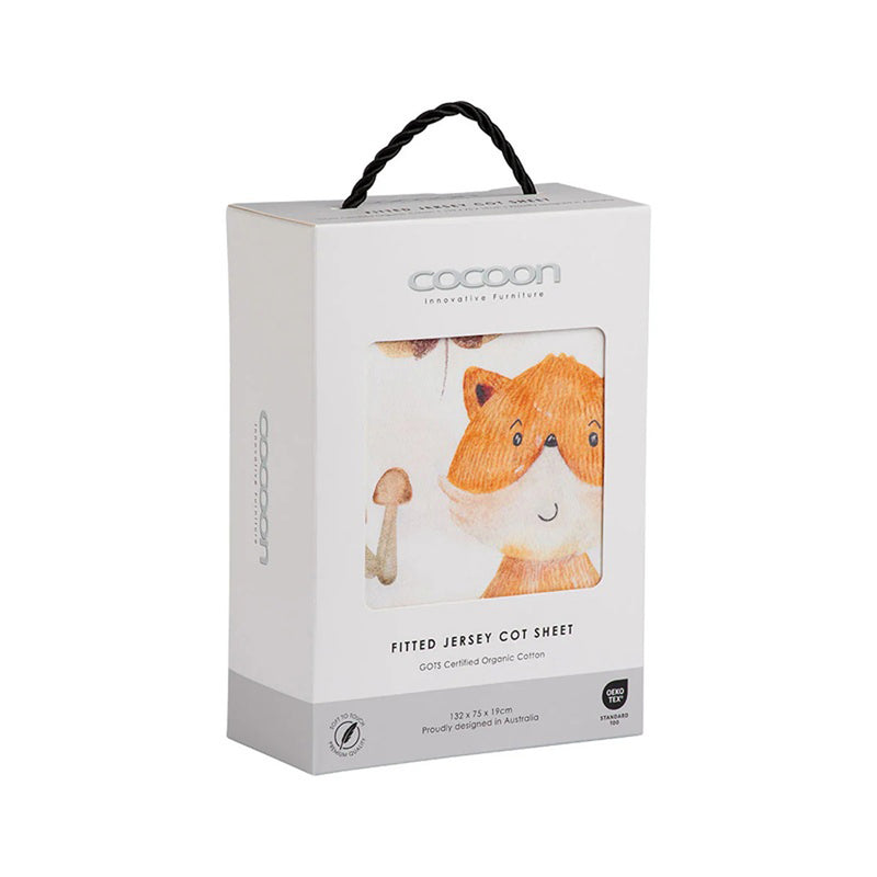 A white box with a black handle showcases the Cocoon Fitted Sheet Cot Forest Friends, featuring a GOTS certified fitted jersey cot sheet with an illustration of a smiling orange fox and mushrooms, visible through a packaging window.