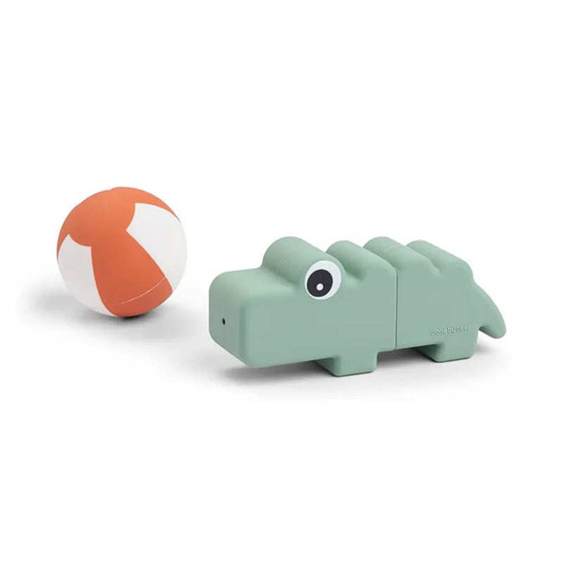 The Done By Deer Bath Squirter Toy Croco Green, featuring a playful silicone crocodile design and large eye, sits beside a small orange and white beach ball—ideal for developing fine motor skills.
