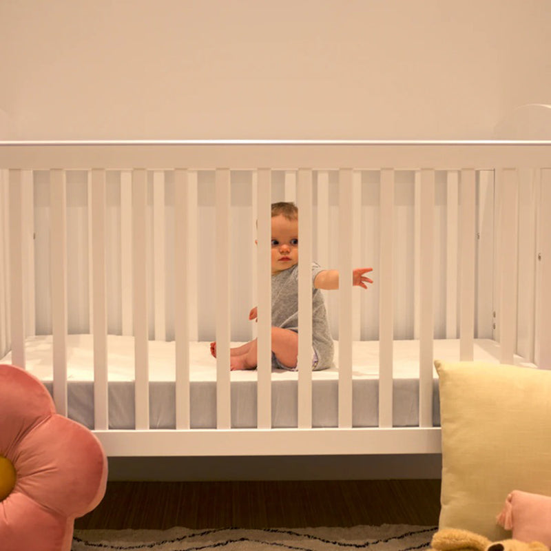 A baby sits in the center of a Grotime Pearl 4 in 1 Cot V2 with a Grotime Comfort Cocoon Mattress. Soft light fills the room, and plush pillows, including a pink flower shape, are visible outside the cot.