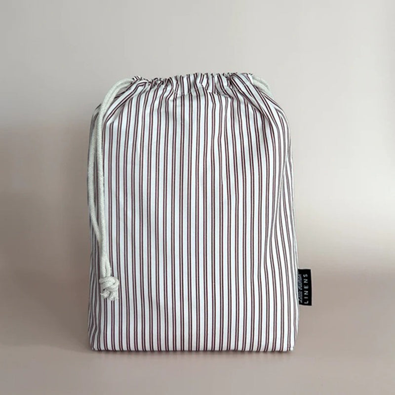 The Little Humans Linens Waterproof Mini Cot Sheet is displayed upright in a drawstring bag with red and white stripes, white drawstrings, and a black tag—ideal for storing your mini cot sheet.