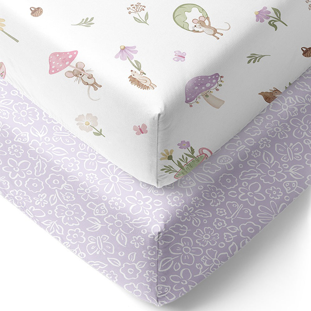 The Living Textiles Cot Fitted Sheet Whimsical Wonderland by Living Textiles includes one white sheet with pastel flowers, mushrooms, mice, and hedgehogs, and one lavender sheet with a white floral pattern—breathable nursery essentials.
