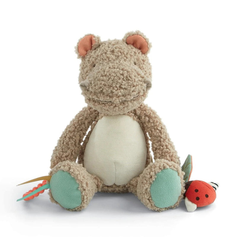 The Mamas & Papas Activity Toy Henry Hippo is a plush sensory toy featuring a beige, curly texture, teal feet, cream belly, and pink inner ears. It sits upright holding a red and green ladybug activity toy and another colorful developmental toy.