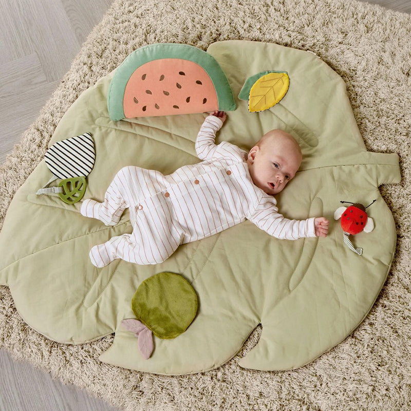 A baby in a striped onesie lies on the Mamas & Papas Playmat Babyplay Leaf by Mamas & Papas, surrounded by soft, colorful toys—like a ladybug, fruit slice, and leaves—perfect for tummy time on a cozy rug.