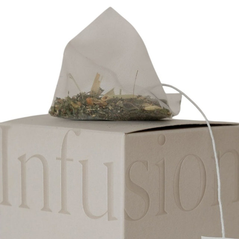 A pyramid-shaped Mammae Bosom Ritual Infusion Nursing Tea Bag filled with organic lactogenic herbs rests atop a beige box, with “Infusion” partially embossed and visible on the packaging.
