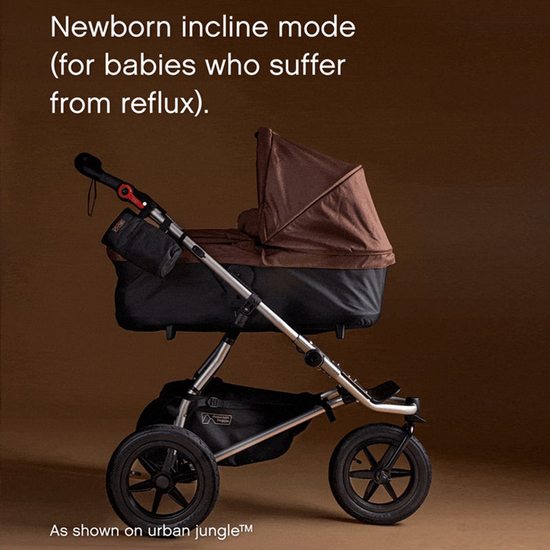 A brown and black Mountain Buggy Carrycot Plus for Terrain V4 shown in incline mode, ideal for newborns with reflux, on a brown background. White text: "Newborn incline mode (for babies who suffer from reflux). As shown on urban jungle™.