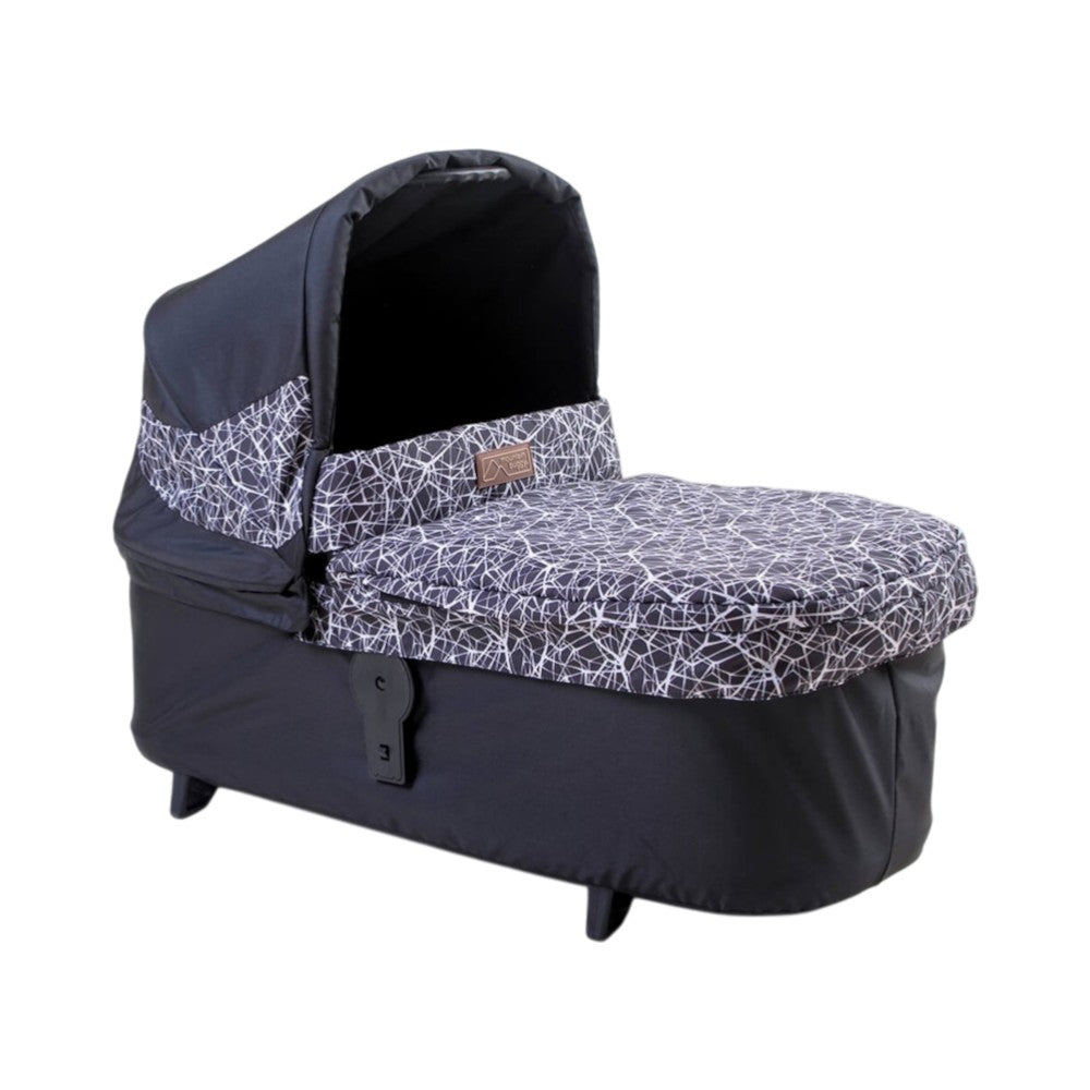 The Mountain Buggy Carrycot Plus for Terrain V4 is a black lie-flat baby bassinet with a hood and white abstract line pattern, elevated on short legs for infants’ comfortable rest.