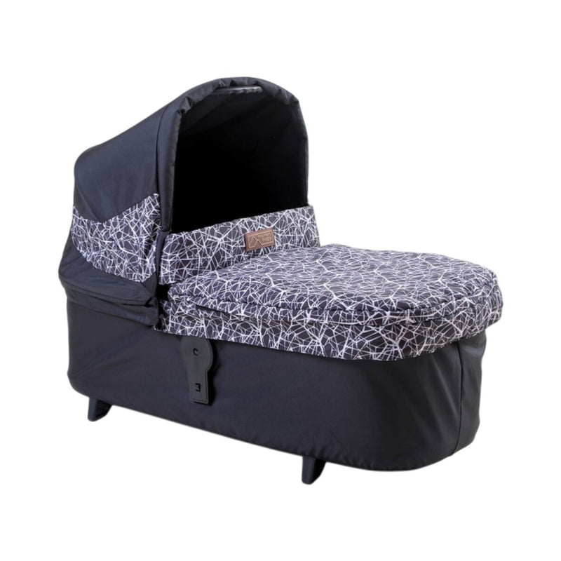 The Mountain Buggy Carrycot Plus for Terrain V4 is a black lie-flat baby bassinet with a hood and white abstract line pattern, elevated on short legs for infants’ comfortable rest.